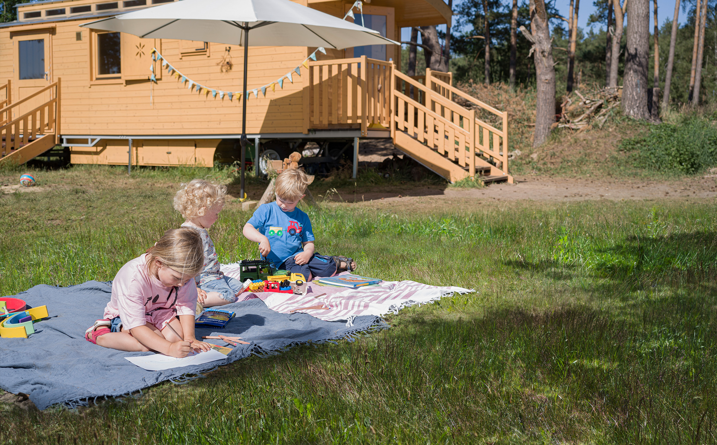 Kinder sitzen im Sommer auf der Wiese und spielen. Im Hintergrund steht ein schöner Bauwagen aus Holz unter Bäumen.